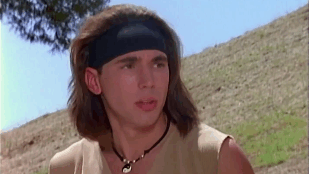 10 Insanely Popular TV Shows From the ‘90s You Can Stream on Netflix 27 Scene from "Mighty Morphin Power Rangers" featuring Tommy Oliver with long hair wearing a black headband in an outdoor setting.