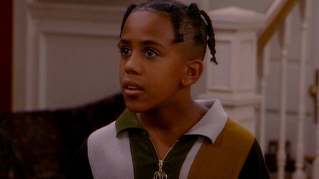 10 Insanely Popular TV Shows From the ‘90s You Can Stream on Netflix 39 Scene from "Sister, Sister" showing Roger with short dreadlocks wearing a white and green striped shirt, looking thoughtful.