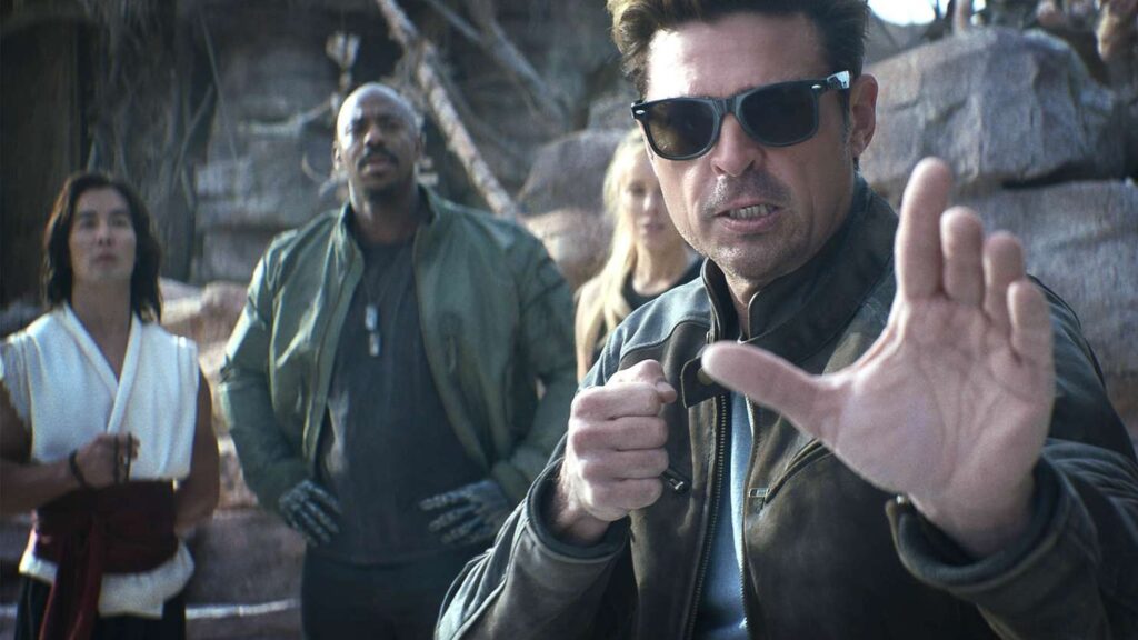 Mortal Kombat 2: Everything We Know So Far 1 Karl Urban as Johnny Cage in Mortal Kombat 2, wearing his signature sunglasses and making a dismissive hand gesture, with other characters visible in the background.