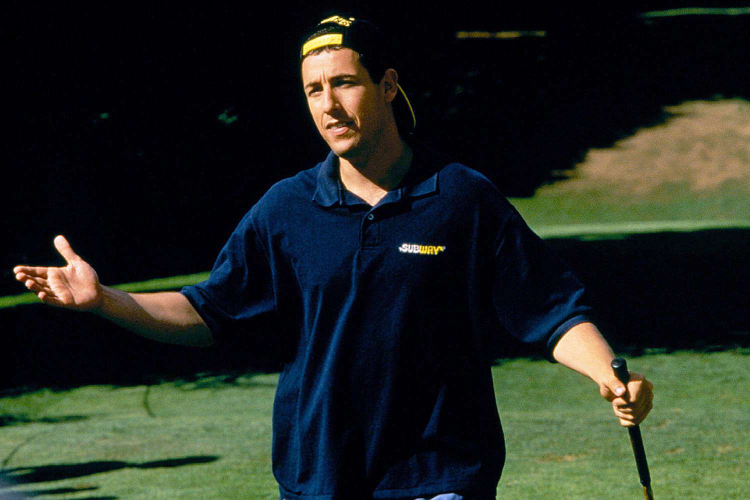 Adam Sandler in a dark blue t-shirt, standing a a field with play stick.