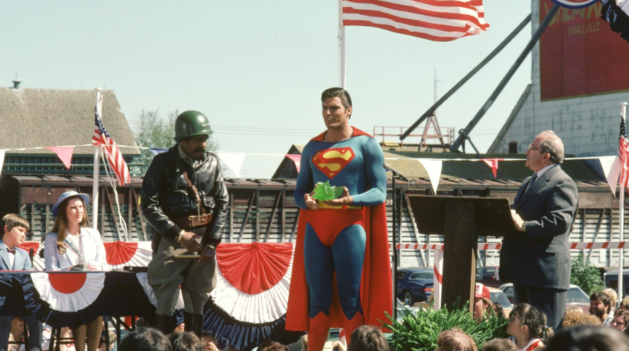 Superman stands on a stage holding a glowing green object, flanked by a man in military attire and another man speaking at a podium, with an American flag flying behind them.