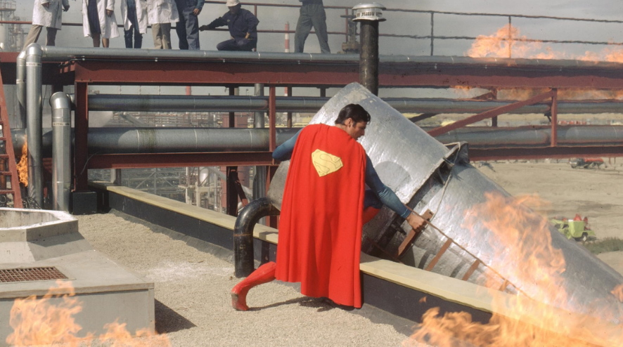 Superman is on a rooftop. He is to lift or move a large piece of debris, with people watching from above.