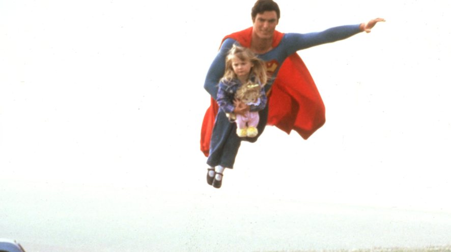 Superman is flying through a bright, clear sky, carrying a young girl in his arms.