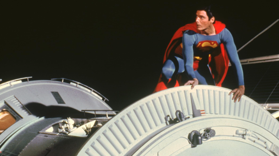Superman is crouching on a space station or large satellite. He is looking to the side.
