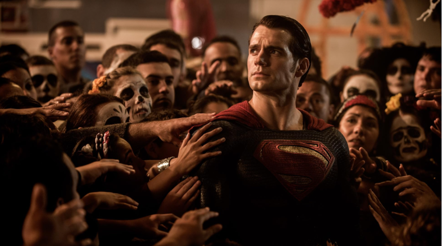 Superman is standing among a crowd of people, some with face paint or masks, reaching out to touch him. But he is looking upward.