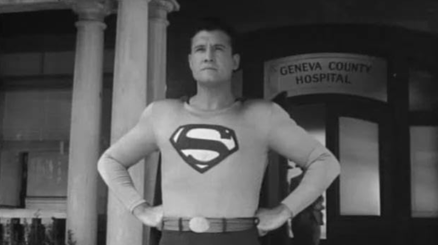 Superman stands with his hands on his hips in front of a building.
