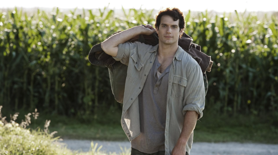 Clark Kent is standing in a field of tall green plants, carrying a jacket and a bag over his shoulder.