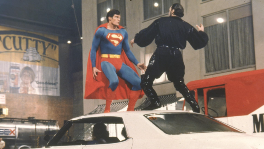 Superman and another man in black are standing on top of a white car.
