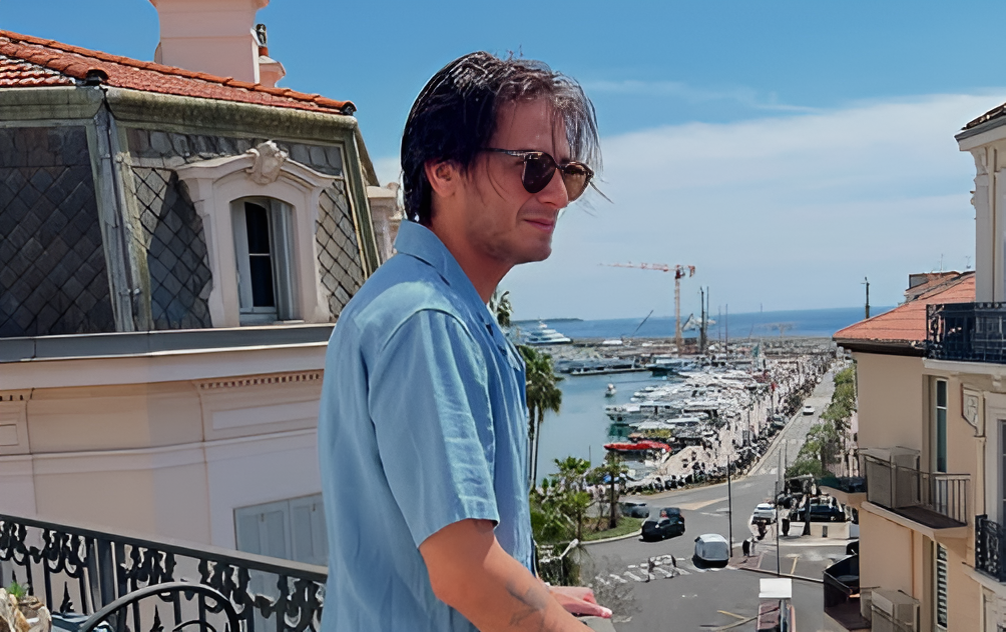 Billie Eilish Relationship History: All Her Boyfriends Before Nat Wolff 2 Matthew Tyler Vorce in a light blue casual shirt and sunglasses standing on a balcony overlooking a Mediterranean coastal town with boats in the harbor and red-tiled roofs.