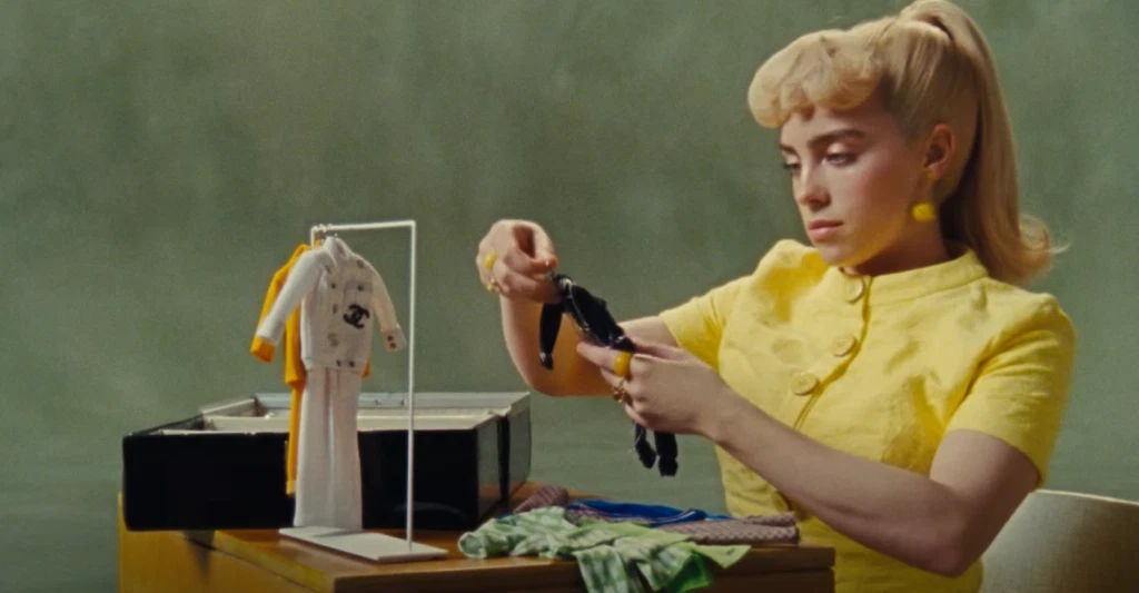 Billie Eilish Relationship History: All Her Boyfriends Before Nat Wolff 4 Billie Eilish, wearing a bright yellow collared shirt, sitting at a table with craft supplies including fabric and tools in the "What Was I Made For" music video.