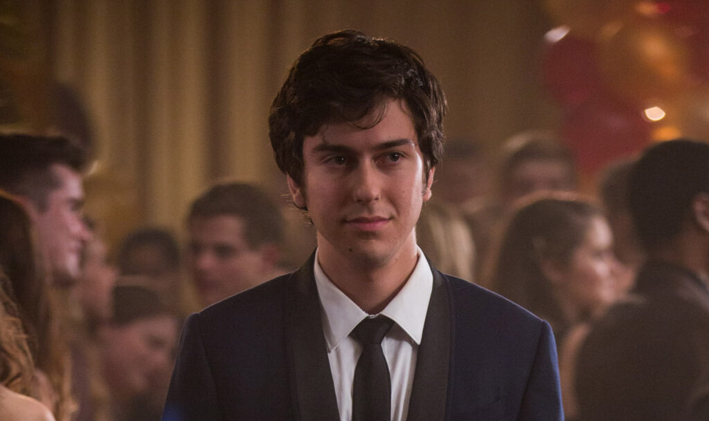 Billie Eilish Relationship History: All Her Boyfriends Before Nat Wolff 5 Nat Wolff wearing a black suit jacket, white dress shirt, and black tie, standing in a formal indoor venue with people in the background in a scene from "Paper Towns."