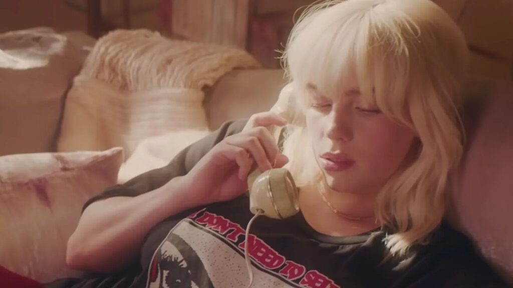 Billie Eilish Relationship History: All Her Boyfriends Before Nat Wolff 6 Billie Eilish sitting casually on a couch, wearing a black graphic t-shirt with red text, in the music video of "Happier Than Ever."