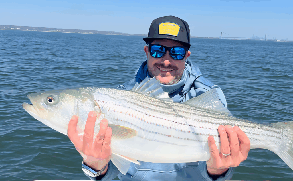 All 21 ‘Top Chef’ Winners and How Their Life Looks Like Today After Winning the Prize Money 1 Harold Dieterle in blue sunglasses and cap holding large striped bass fish while on a boat, with ocean and distant shoreline in background.