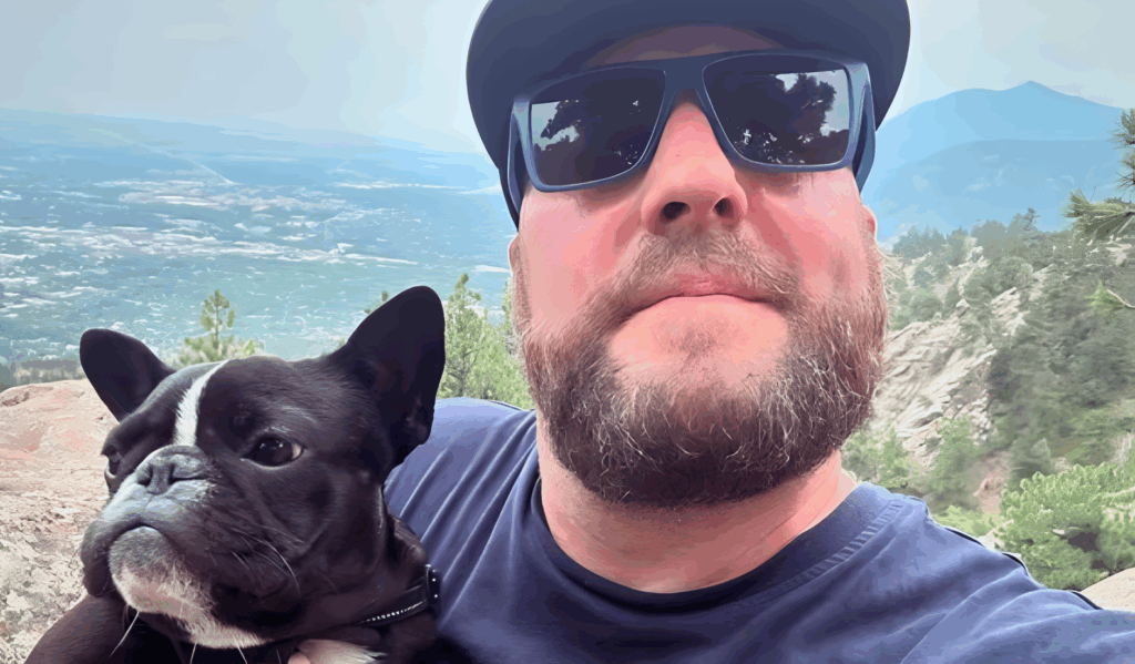 All 21 ‘Top Chef’ Winners and How Their Life Looks Like Today After Winning the Prize Money 5 Hosea Rosenberg in black cap and sunglasses taking selfie with his French Bulldog, with scenic mountain valley landscape in background.