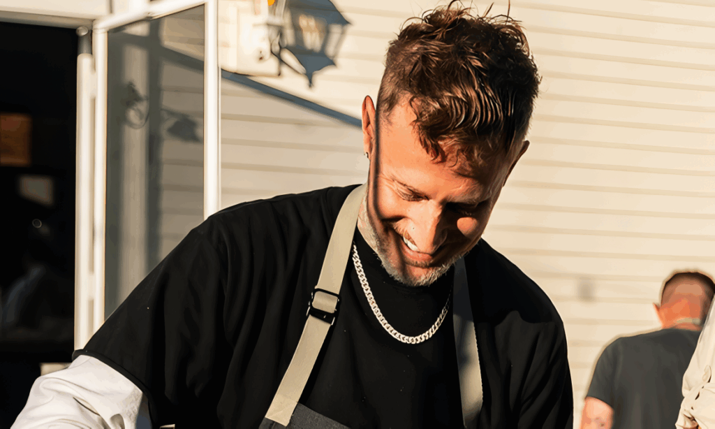 All 21 ‘Top Chef’ Winners and How Their Life Looks Like Today After Winning the Prize Money 6 Michael Voltaggio in black shirt and tan apron, smiling while working outdoors with white buildings in background.