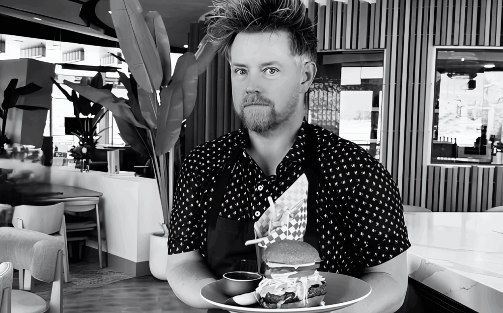 All 21 ‘Top Chef’ Winners and How Their Life Looks Like Today After Winning the Prize Money 8 Black and white photo of Richard Blais in polka dot shirt standing in a restaurant with a gourmet burger and fries, with modern restaurant interior in background.