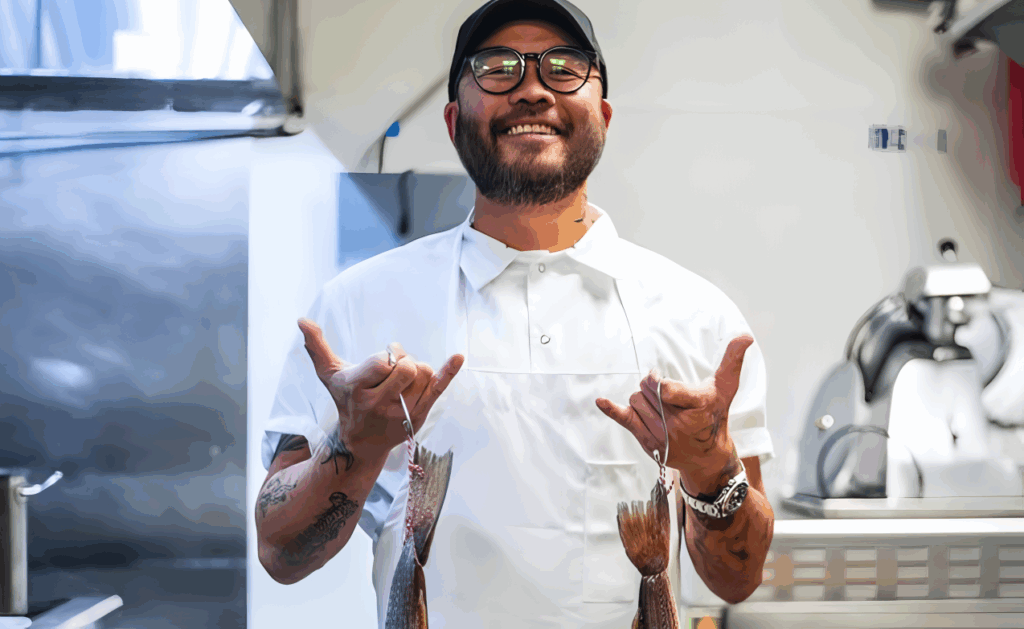All 21 ‘Top Chef’ Winners and How Their Life Looks Like Today After Winning the Prize Money 9 Paul Qui with glasses and black cap in white chef's coat, holding a fish in each hand by its hook while standing in a professional kitchen.