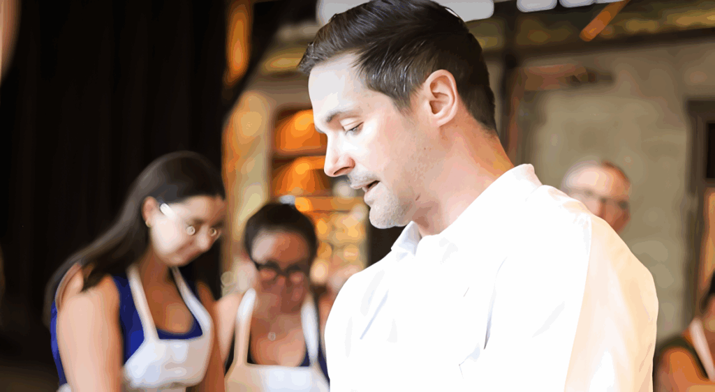 All 21 ‘Top Chef’ Winners and How Their Life Looks Like Today After Winning the Prize Money 11 Nicholas Elmi in white chef's coat speaking or presenting in a restaurant kitchen, with other chefs and staff visible in the blurred background.