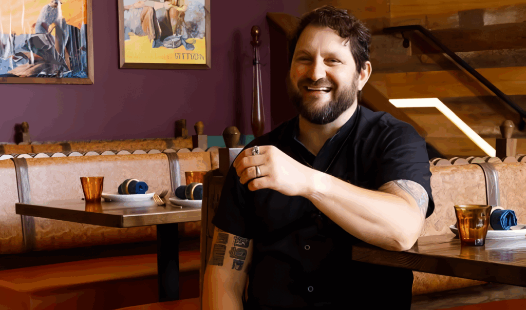 All 21 ‘Top Chef’ Winners and How Their Life Looks Like Today After Winning the Prize Money 18 Gabriel "Gabe" Erales in black chef's coat sitting at a restaurant table with colorful artwork on purple walls in the background.