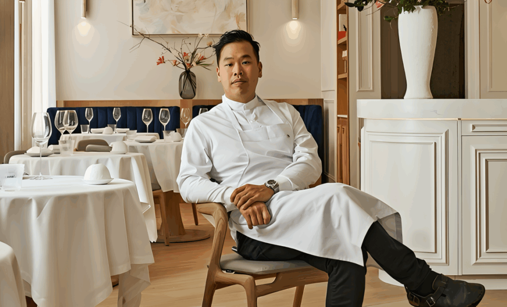 All 21 ‘Top Chef’ Winners and How Their Life Looks Like Today After Winning the Prize Money 19 Buddha Lo in crisp white chef's coat sitting confidently in an upscale restaurant dining room with white tablecloths and elegant decor.