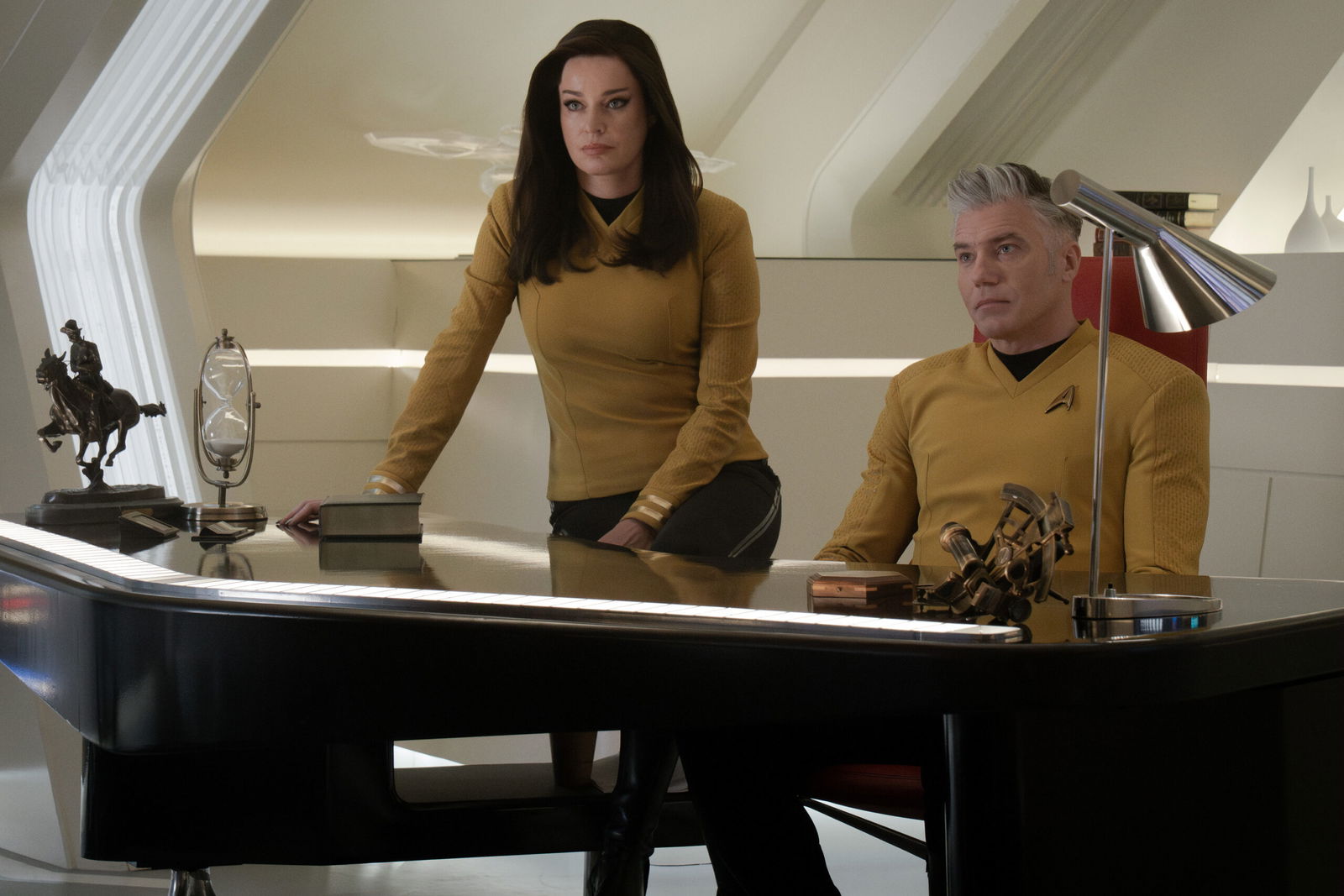 A still from Star Trek: Strange New Worlds Season 3 featuring Una and Pike standing behind and sitting at a desk respectively.