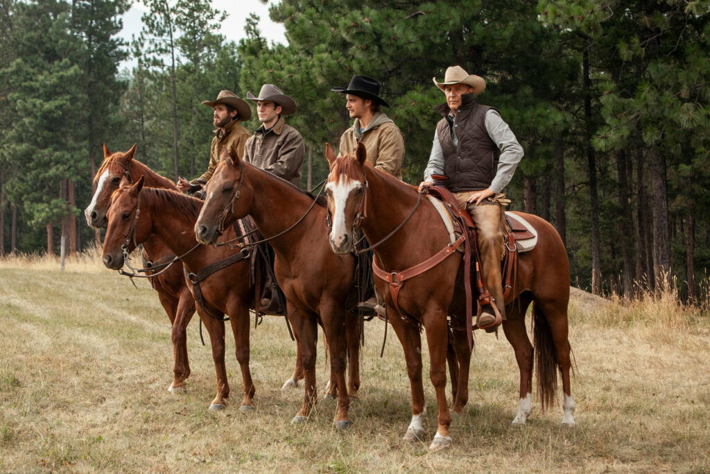 The cast of Yellowstone sitting atop horses and looking in a still from the show.