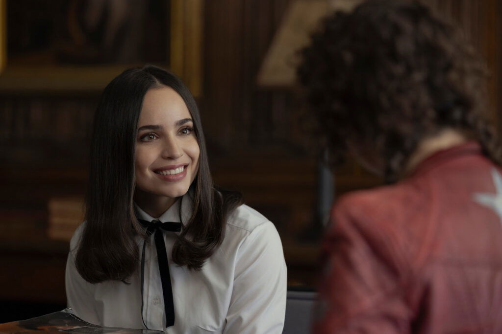 My Oxford Year Might Be the Emotional Cousin of Emilia Clarke’s Saddest Movie 4 Sofia Carson smiling with straight hair, wearing a white shirt with black bows in front of someone wearing a red jacket.