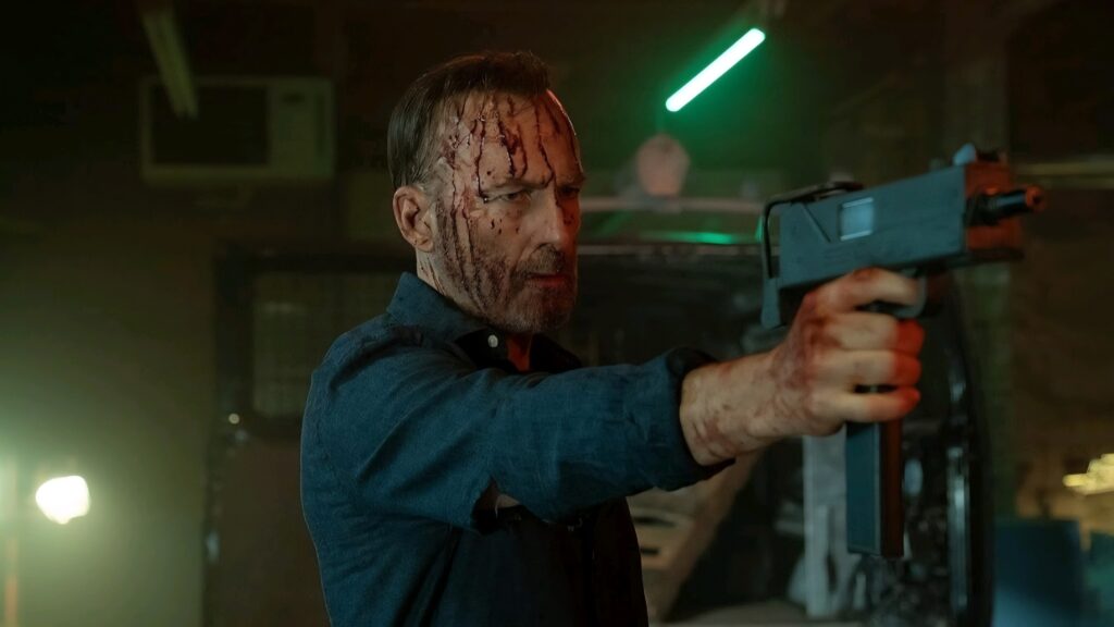 Bob Odenkirk looking tired, bleeding from his head, and pointing a gun