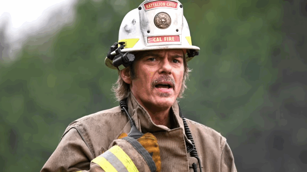 Billy Burke is wearing a "BATTALION CHIEF" helmet and he is speaking, holding a radio. He is giving instructions it seems.