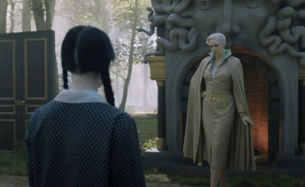 Larissa wearing a cape and a long dress, standing in front of Wednesday. 