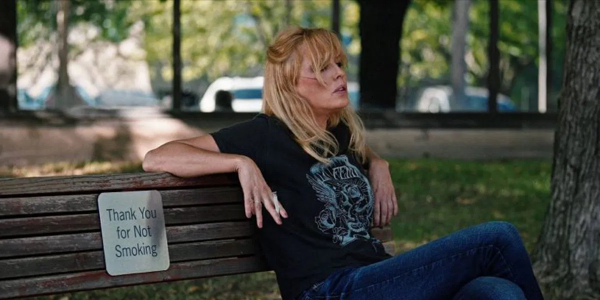 Kelly Reilly as Beth Dutton in Yellowstone sitting on a park bench and smoking even though there's a sign saying not to smoke