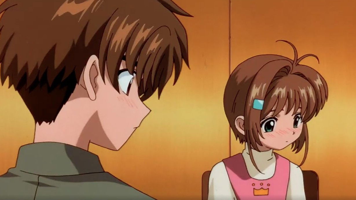 Two young people in an indoor setting with warm lighting. Syaoran Li with brown hair facing toward Sakura Kinomoto with lighter hair wearing a white top with a small crown design.