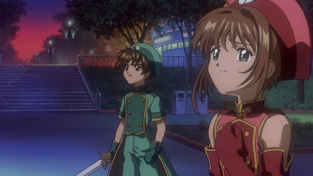 Two characters in elaborate costumes in an evening park setting. Syaoran Li in a teal military-style uniform in the background, Sakura Kinomoto in a red dress-like outfit in the foreground.