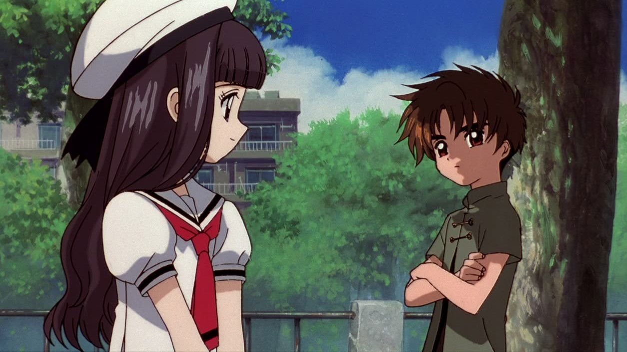 Tomoyo Daidouji with dark hair in a school uniform with a red tie standing outdoors, with Syaoran Li in casual clothes visible in the background.