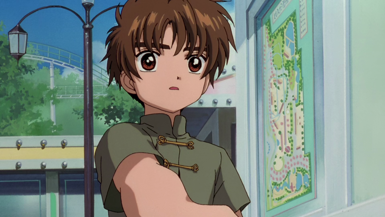 Syaoran Li with brown hair in a green outfit with decorative buttons, shown outdoors with arms crossed in a confident pose.