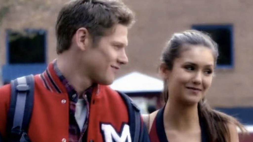 Matt in his high school letterman jacket standing with Elena who is smiling, both appearing to be teenagers during an earlier season of "The Vampire Diaries."