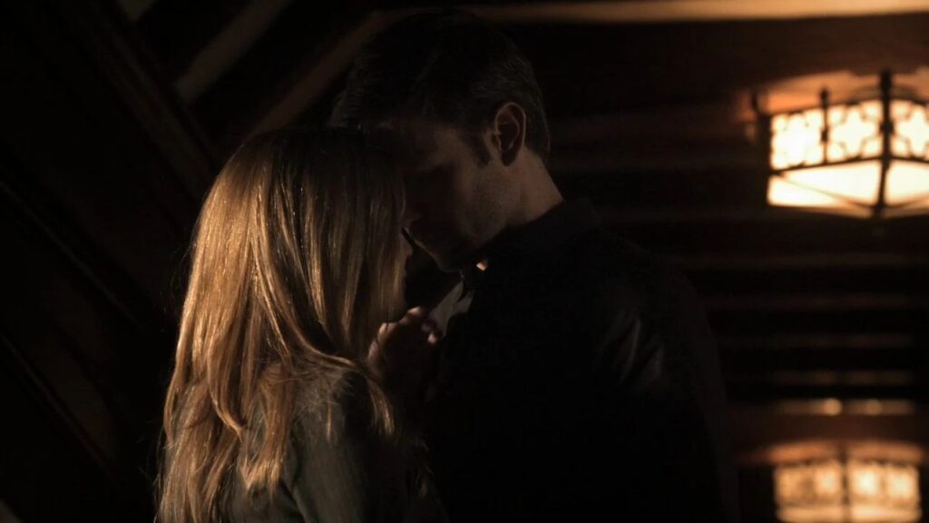 Alaric and Jenna in an intimate moment indoors with dramatic lighting, appearing very close together.