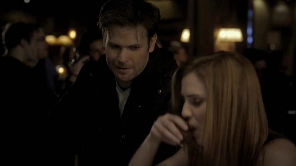 Alaric standing behind Jenna in a dimly lit indoor setting. Jenna is having a drink and Alaric is looking at her.