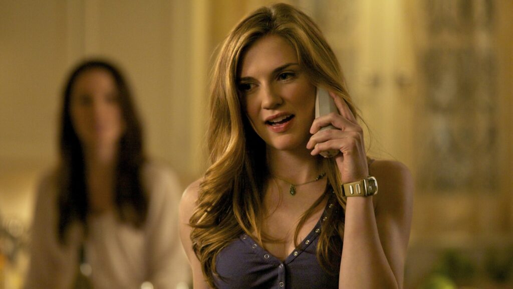 Jenna talking on a phone while standing indoors, wearing a casual shirt and appearing focused on her conversation.