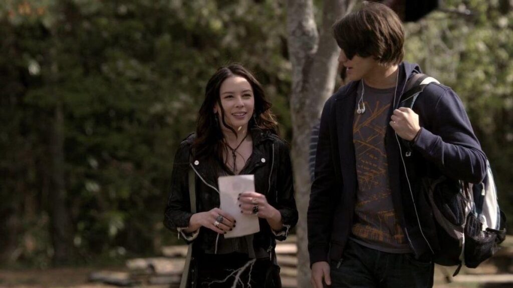 Anna and Jeremy walking together outdoors in a wooded area, with Anna in a leather jacket and Jeremy in casual clothing, both appearing happy.