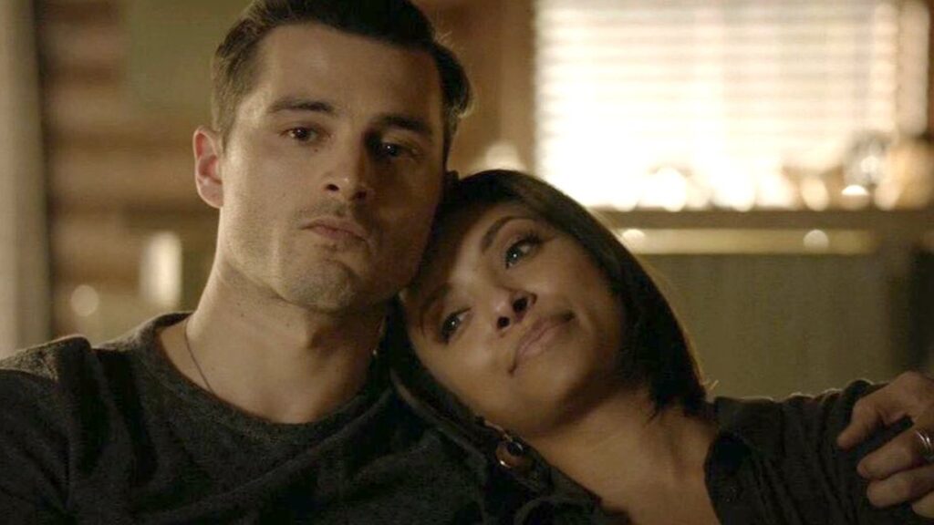 Bonnie and Enzo sitting close together indoors, both wearing dark sweaters and looking content in each other's company.