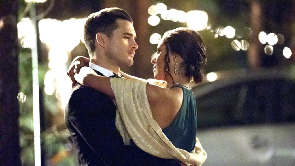 Bonnie and Enzo dancing together at a formal event, with Bonnie in an elegant gown and Enzo in a tuxedo, surrounded by warm string lights in the background.