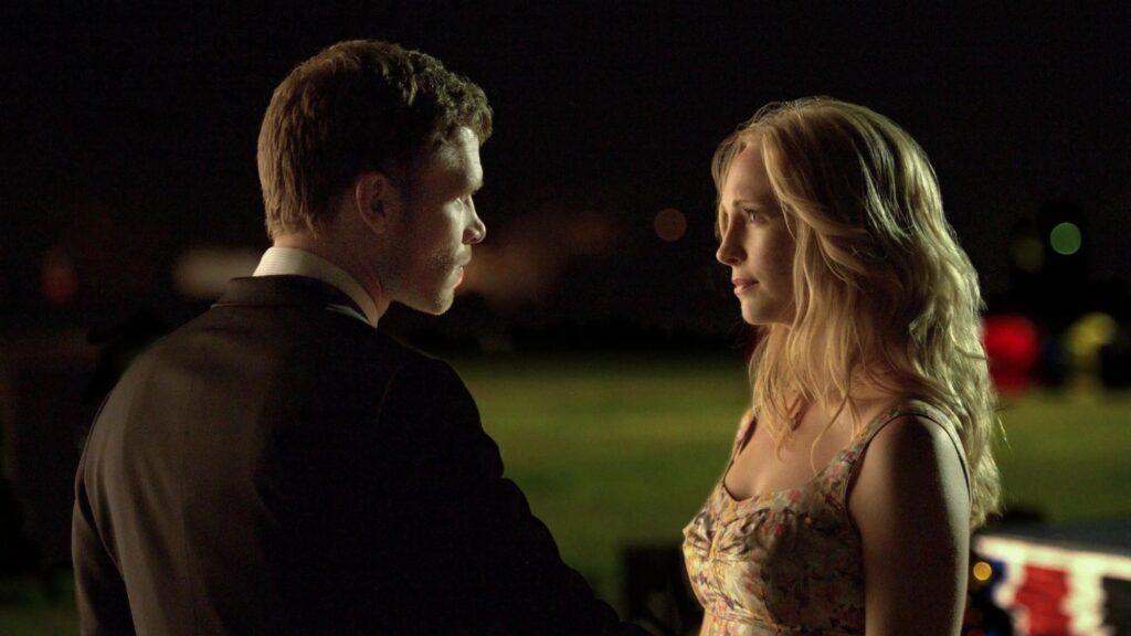Caroline and Klaus at a formal event, both dressed elegantly and standing close together during a night-time scene.