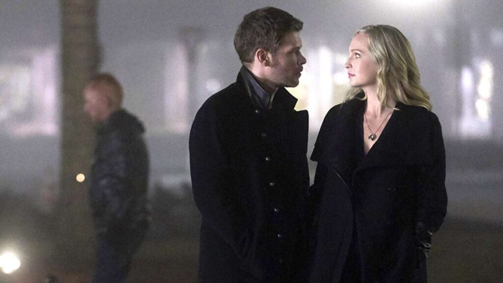 Klaus and Caroline in a nighttime scene outdoors, both in dark clothing with atmospheric lighting.