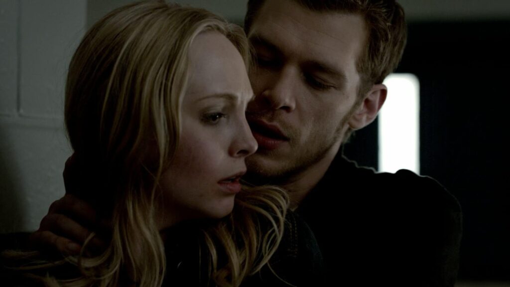 Klaus and Caroline in an intimate embrace indoors, with Klaus holding Caroline close in dramatic lighting.