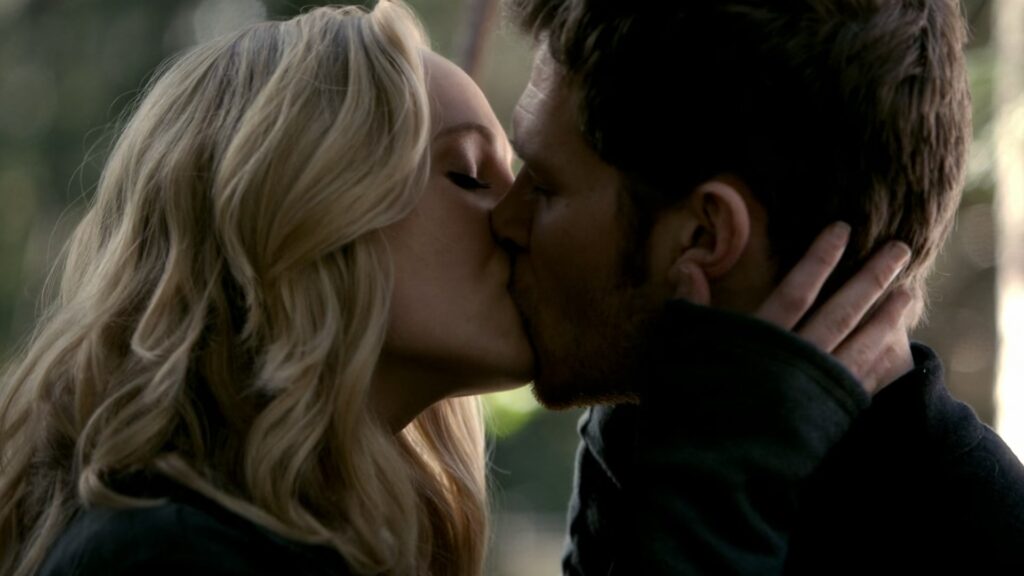 Caroline and Klaus sharing a passionate kiss outdoors in a wooded setting with natural lighting.