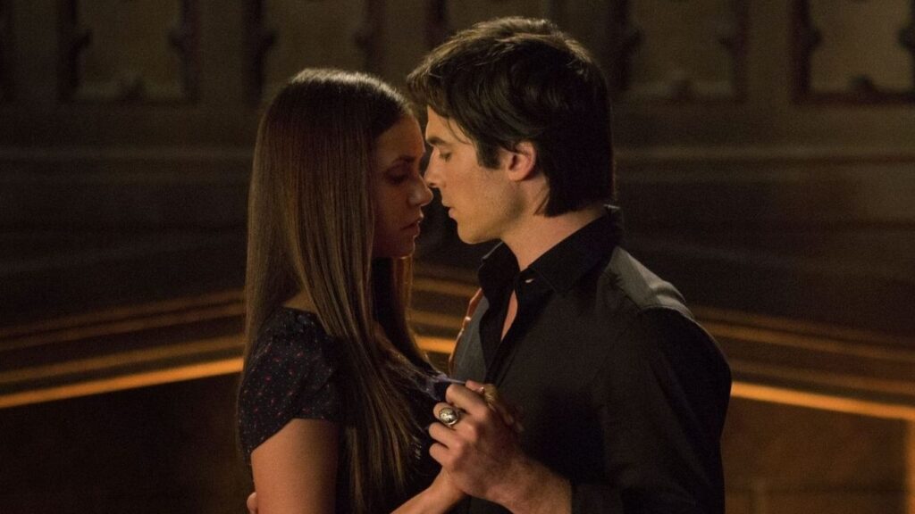 Elena and Damon in an intimate scene, standing close together with warm lighting highlighting their faces and slow-dancing.