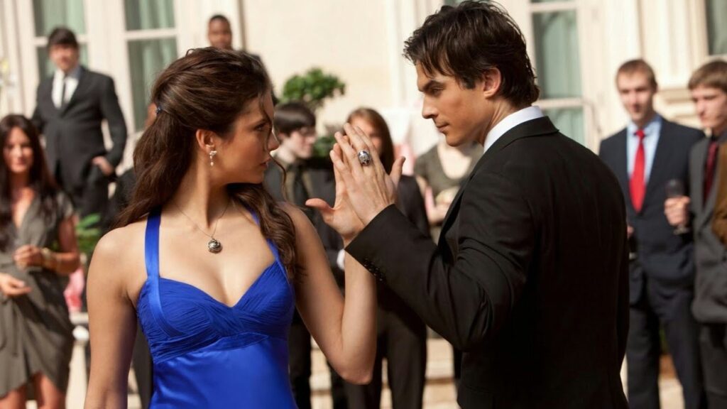 Elena and Damon at a formal dance, with Elena in a blue dress and Damon in a suit, their hands touching. Other people are visible in the background.