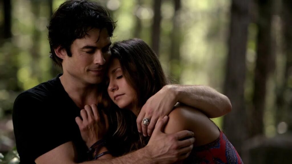 Damon and Elena embracing each other in a wooded outdoor setting, both appearing peaceful and content.