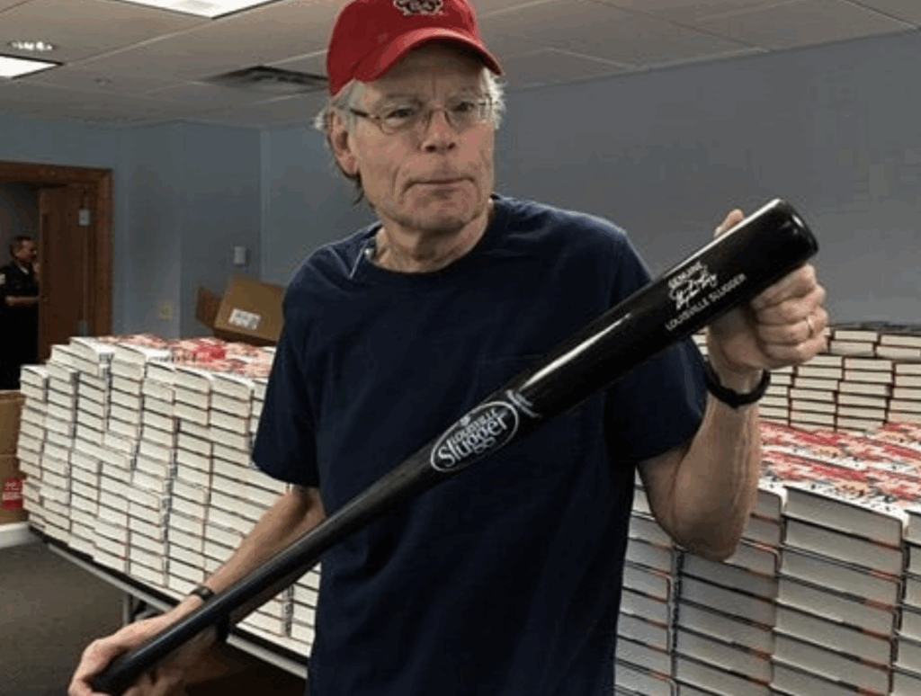 Stephen King wearing a red cap and holding a baseball bat.